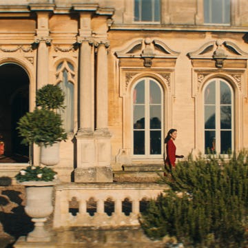 A woman walks near a grand mansion with ornate architecture.