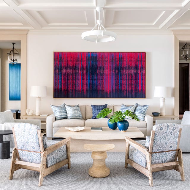 a living room with a off white sofa with a large blue and red abstract painting on the wall behind and wood and upholstered chairs
