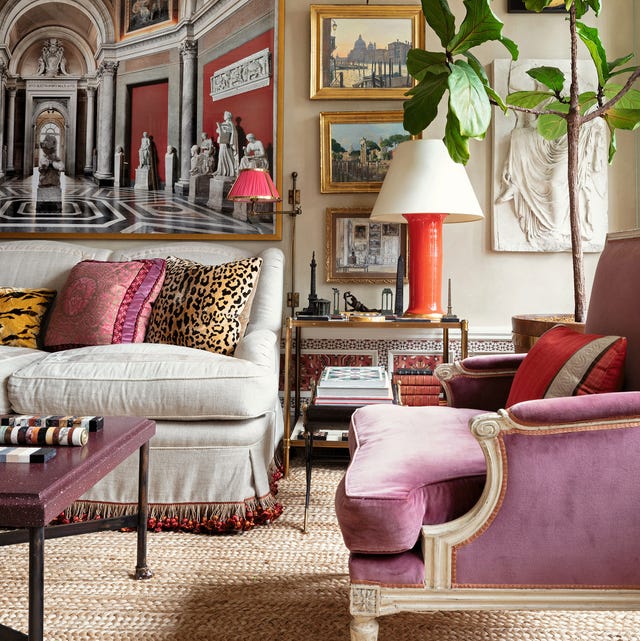 elegant living room with a plush linen sofa with fringe on the skirt and a pink velvet upholstered regency chair in the foreground and classical artwork in gold frames on the back wall and a side table with books and an modernistic orange ceramic base lamp and a jute rug on the floor