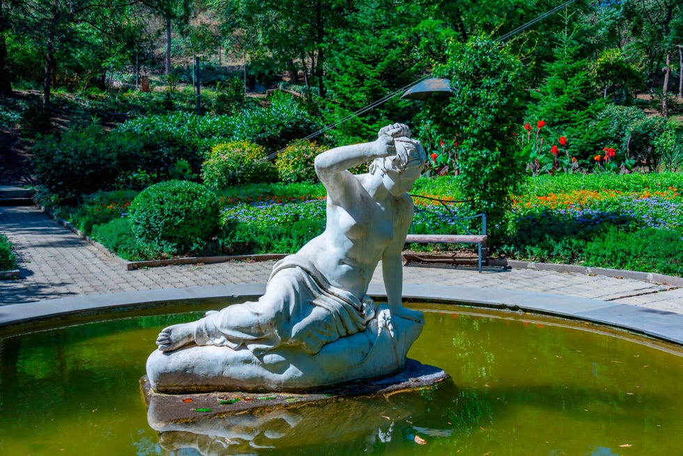 Sculpture of a reclining figure in a pond surrounded by greenery and flowers. Sculpture of a reclining figure in a pond surrounded by greenery and flowers.
