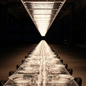 A long, elegantly set dining table illuminated by overhead lights. A long, elegantly set dining table illuminated by overhead lights.