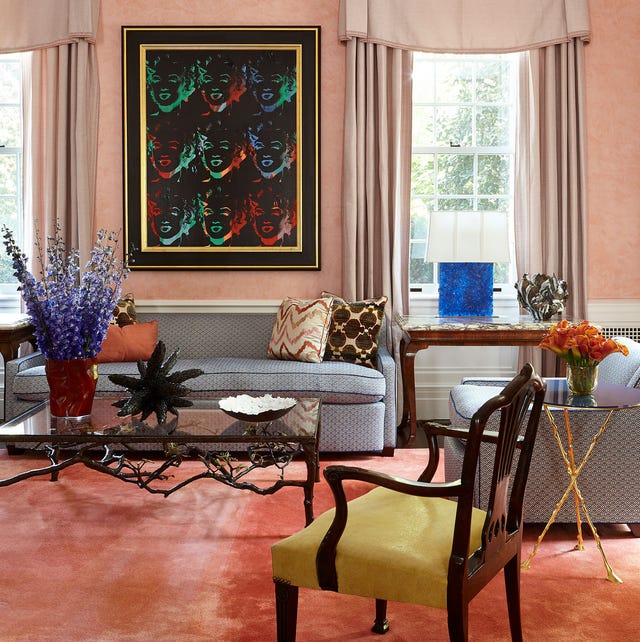 a pink painted living room with orange velvety rug a gray sofa with blue trim and matching chair and a dark wood armchair with yellow seat and floor to ceiling lighter pink curtains and a large dark framed artwork over the sofa and between the windows