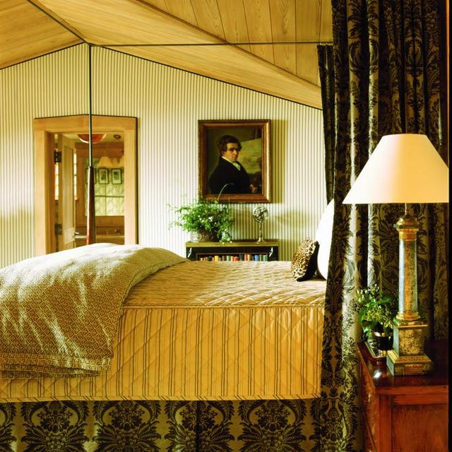 a colonial looking bedroom with a four poster bed with green patterened curtains and matching bed skirt and a yellow quilted blanket and a portrait on the far wall and a lamp