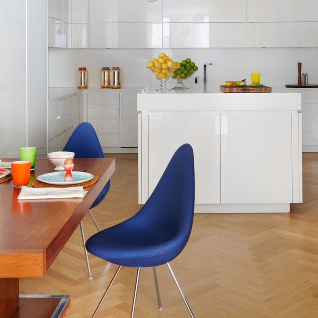 a kitchen with a honey colored table and blue chairs and a white lacquer counter with sink and cabinets at back and a light chevron pattern floor