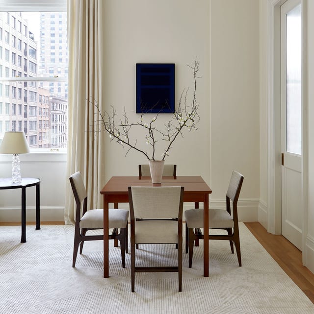 small wood table with a vase with branches on int and with linen and wood chairs and a off white rug in a off white room with a window and floor to ceiling curtains