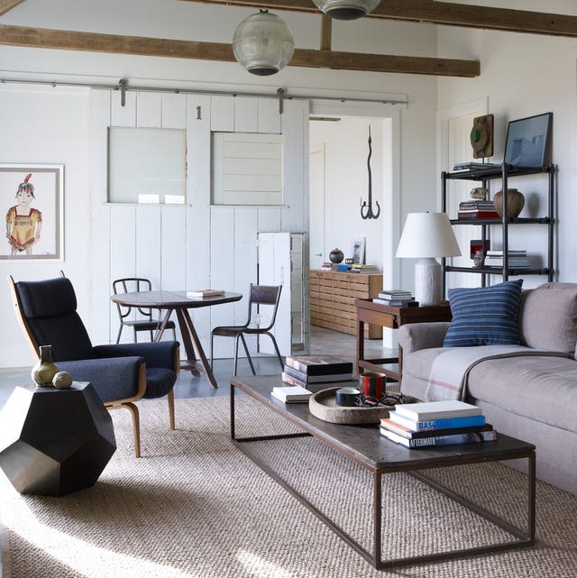 a white painted rustic living room with oval drop lights and a sisal carpet and a rectangular cocktail table and velvety grayish sofa and a dark blue chair with wood accents to the right and in the background a simple tripod table and chairs and an etagere with objects in the corner