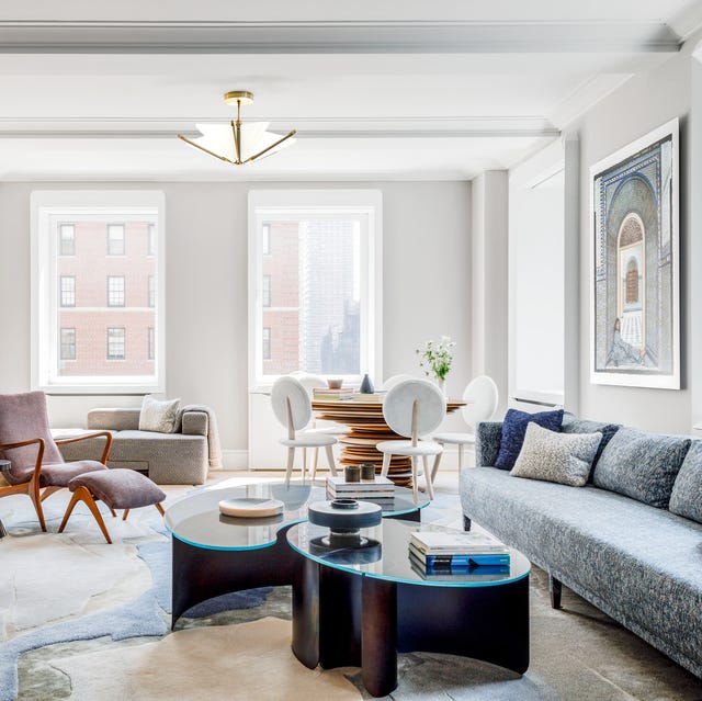 spacious airy living room with long blue upholstered sofa against the right wall and a cocktail table with interlocking sections topped with glass and a rug that looks like colored rock and chairs along the other side