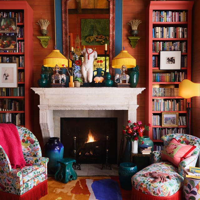 very cozy closeup of a sitting area with red plank walls and a white marble fireplace with a large blue and red mirror over it and jade colored objects and elephant lamps with matching yellow fringe shades and in front of the fire is a small colorful chair and matching chaise with red fringe all on a multicolored carpet and two tall bookcases on either side of the fireplace also painted red