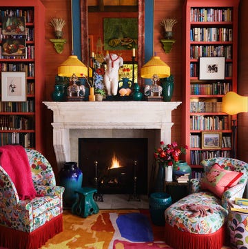 very cozy closeup of a sitting area with red plank walls and a white marble fireplace with a large blue and red mirror over it and jade colored objects and elephant lamps with matching yellow fringe shades and in front of the fire is a small colorful chair and matching chaise with red fringe all on a multicolored carpet and two tall bookcases on either side of the fireplace also painted red