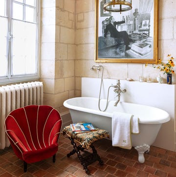 stylish stone wall and terra cotta floor bathroom featuring a vintage footed bathtub a red shell shaped chair and a large framed black and white photograph