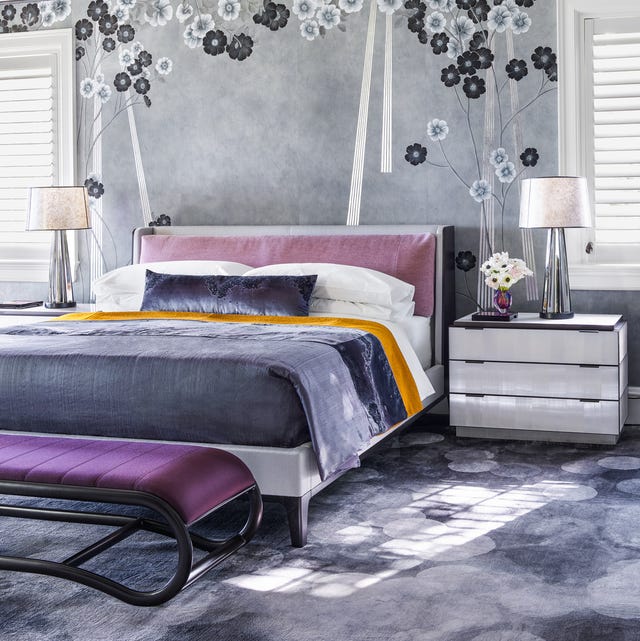 bedroom in soft slate blue and lilac colors with a mottled slate blue carpet and a similar darker colored bedcover and a pink velvety insert into the headboard and a dark purple bench with black bottom at the foot and a white three drawer nightstand and the wall has a wallcovering in gray with black and white simple flowers on parts of it