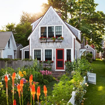the exterior of a cedar shingled two story cottage with window boxes with red flowers, a red screen door, and brick steps, a white picket fence with roses surrounds tall orange flowers, yellow day lilies, pink foxglove