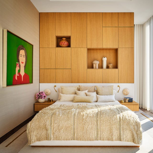 an airy high ceilinged bedroom with a bed and light wood paneled wall with some inset shelves and cabinetry and on the right is a large painting with a green background of a woman in red holding a glass of wine