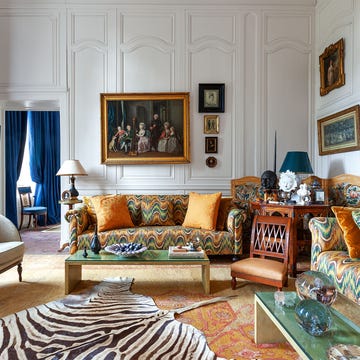 a great room with numerous framed artworks on the paneled walls, two sofas with a swirly colorful print, a wooden chair and an armchair, green glass topped cocktail tables, zebra print rug, deep blue curtains