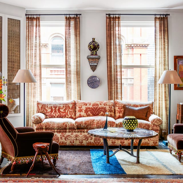 living room with tall curtained windows at back and matching standing lamps on either side of a plush cushy sofa with red and off white pattern upholstery that matches the curtains and a round coffee table and a dark brown velvet chair at left that has tassels along the trim and a trileg side table next to you
