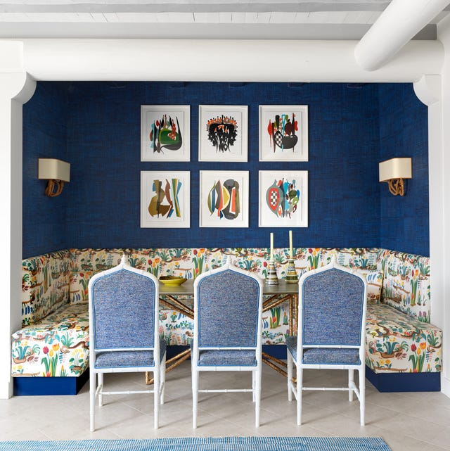 a happy flower printed banquette that wraps around three dark blue walls with six white framed artworks in a grid and tall table at center and three blue chambrey like upholstered chairs pulled up to it and