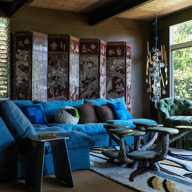 moody living space with bright blue velvety small sectional sofa and a cocktail table with several arms with tops and a large screen against the wall behind the sofa