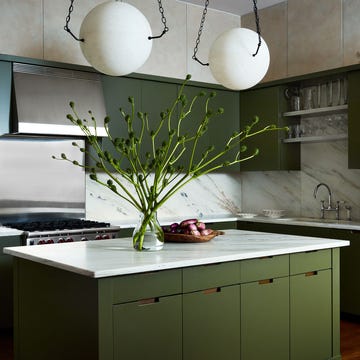 a kitchen has an island with a marble surface and dark olive green cabinets, two alabaster globe pendants suspended by chains, marble backsplashes, stainless oven, sink with open shelving and glasses above