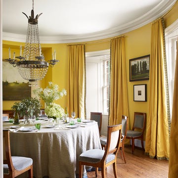 round yellow dining room with wood floors a chandelier and chairs and table with floor length tablecloth and yellow curtains on tall windows