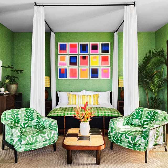 a bedroom with lots of green including a canopy bed with white sheers and a green check blanket and two palm tree patterned green and white armchairs at the foot and a sisal carpet on wood floors and a colorful array of frames over the bed