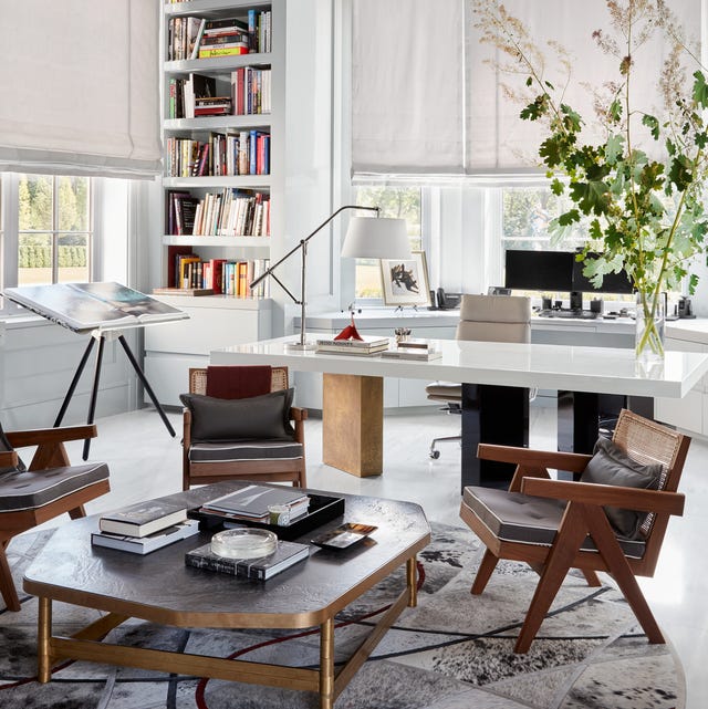 study or den with long white shiny lacquer tri pedestal table with a midcentury low cocktail table and chair over a geometric black and white rug and an easel with flat open book at back and a white inset bookshelf