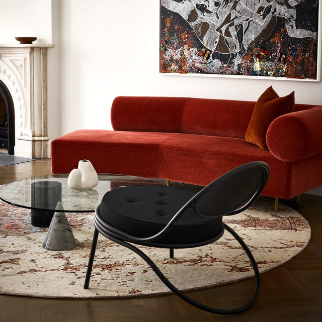 swishy reddish orange velvet armless sofa in a living area with a round rug that looks like a tortilla and a black wire chair and glass cocktail table with three different shaped legs and a large artwork in complementary colors on the wall