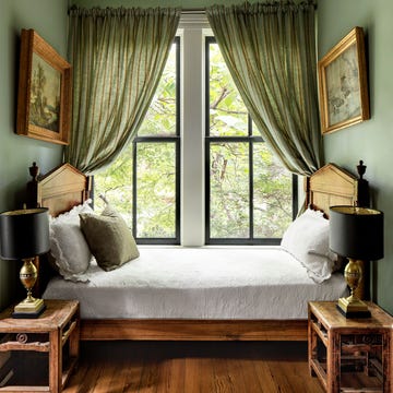tucked into a curtained window nook is a fruitwood daybed with turned urn finials and decorative pillows, two antique chinese side tables each with a bronze lamp, and framed landscape paintings on facing walls