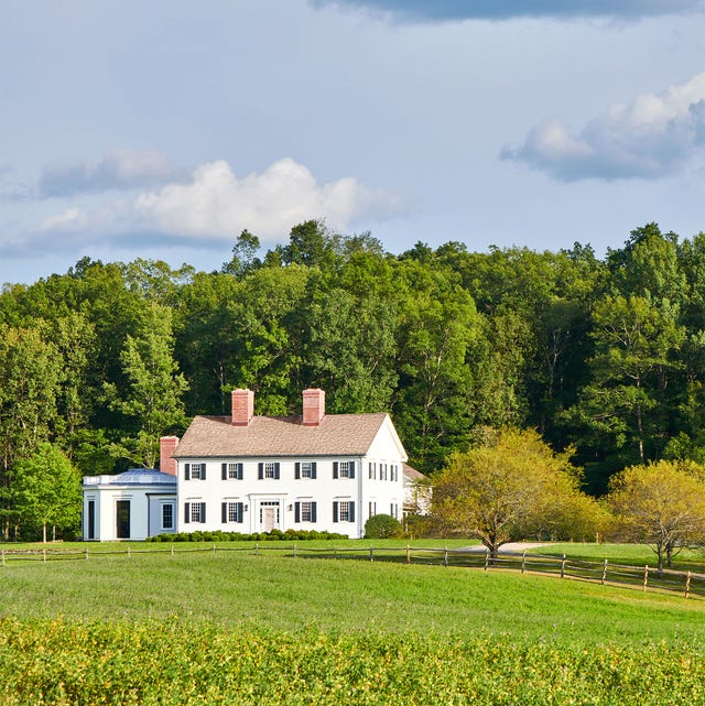a colonial style white house with two floors and black shuttered windows with an octagonal single floor addition sits on a property with an expansive front lawn with a rustic horse fence, and tall trees behind the home