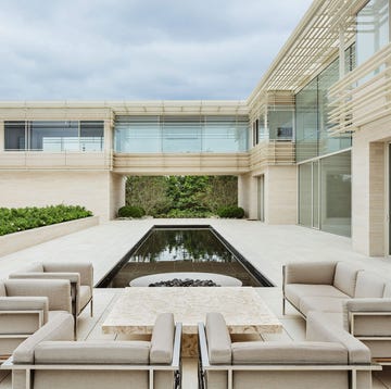 a two level house consisting of two wings, concrete walls and horizontal rails, lots of windows, a terrace with chairs and cocktail table, reflecting pool with a firepit at one end