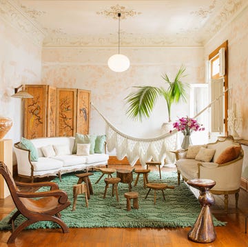 open airy room with a green shaggy rug at center and a series of different shaped wood stools  at center and a white sofa at an angle on the far left and a settee on the right behind which is a tall piece of white column art and opposite are a tall wooden pedestal and a short one with pieces of art on them and a fringed hammock is hung between the two walls at the back behind the furniture
