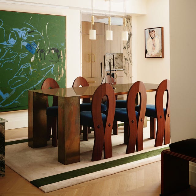 a high glossy wood dining table with chair pulled up to it with ankh shapped backs and blue seats on a cream rug with two green stripes and a large green splatter painting on the wall and glass cabinetry