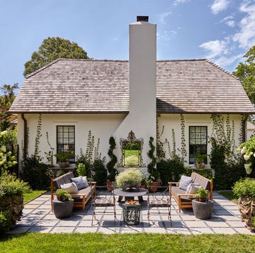 a cottage painted a light blush pink, chimney with a decorative mirror hanging on it, terrace with wood framed sofas with cushions, wrought iron side chairs and table, concrete tiles, flowering plants, grass lawn