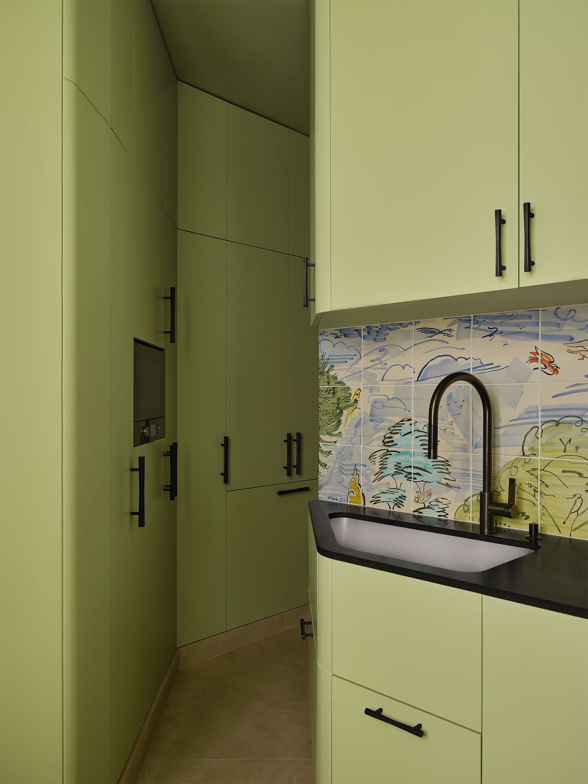 a kitchen has light avocado colored cabinets with black pulls along a corridor and above and below a sink set in a black coun
