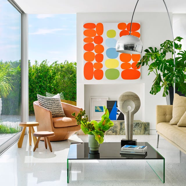 spare midcentury modern styled room with glass walls and white shiny floors and a light brown swivel chair and some low three legged wood tables and a white fireplace with a painting overhead that has explosive orange accents and a settee to the right and a simple low table with objects and plants on it