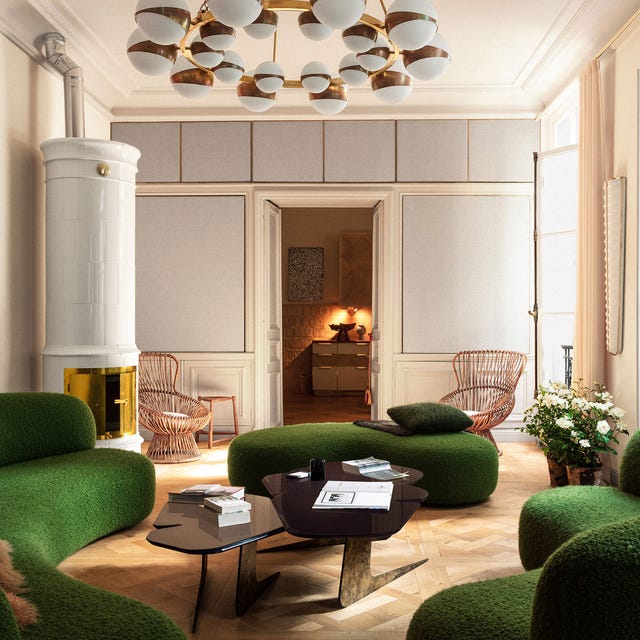 a classic living room with curvy green popcorn fabric sofas and matching chaises and a funky black cocktail table with l shaped legs in a metal finish and a white and metal band chanelier at top and on the left a tall white ceramic fireplace stove with metal doors