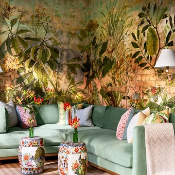 a seating area with a sea foam colored velvet sectional sofa with accent pillows, colorful drumlike tables, two sconces with white shades, walls covered in a wild multicolored print with large botanicals