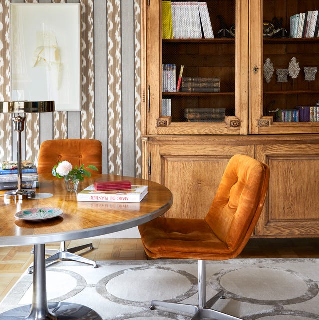 seating area with round pedestal table and two orange velvet armless chairs on swivels and a large oak cabinet with glass doors behind it and a white on white rug with round designs on the floor