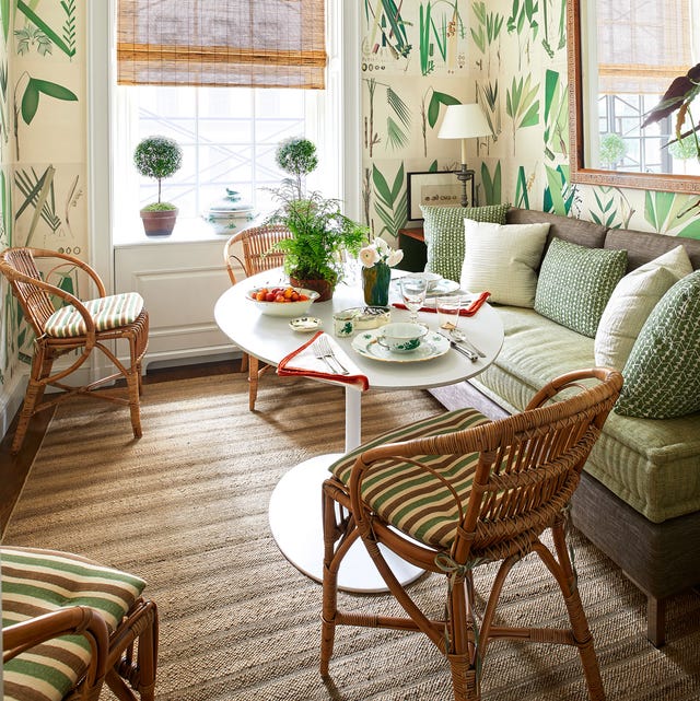 a cozy seating area with white pedestal table set with breakfast plates and cups and a leafy wallpaper and rattan chairs with striped pillow seats and a sofa with platform and velvety light green cushion
