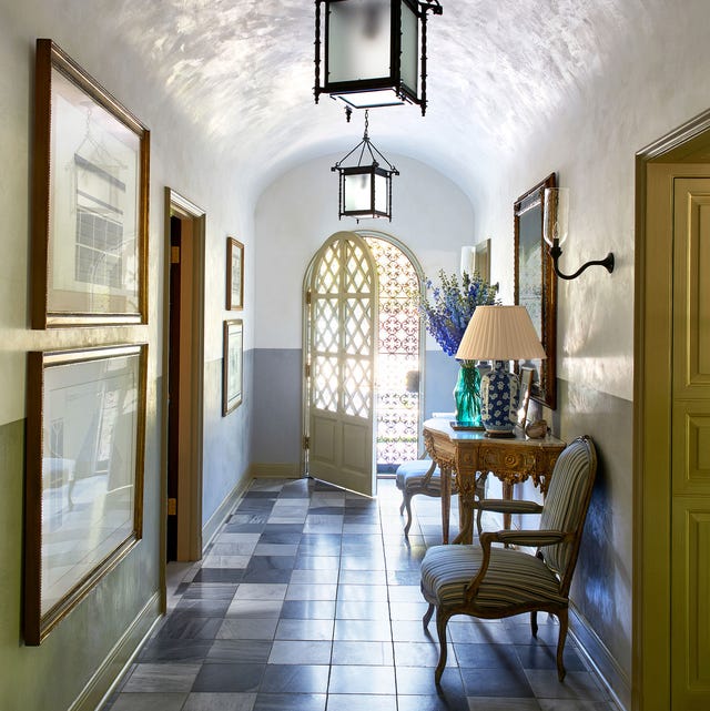 entry hall with variegated gray tile and artwork on the walls and a gilded demilune table with armchairs flanking it and an oval arched door with screen at the far end and lantern hanging overhead