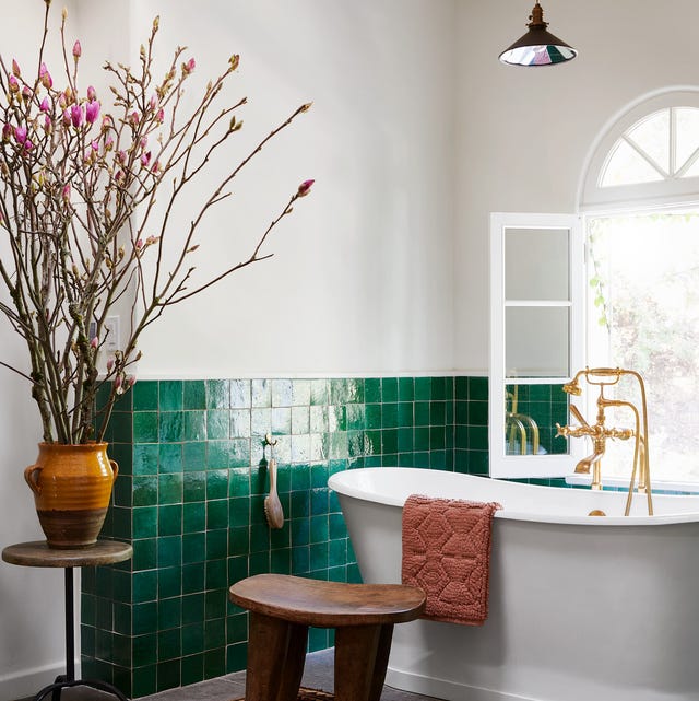 bathroom with half green shiny tile and standalone tub with gold faucets and a wood low stool next to it and a red patterened rug and a window at back with a view