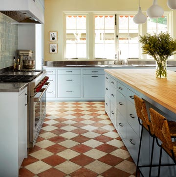 kitchen with light blue cabinetry, light yellow walls, island with butcher block top and two rattan backed stools and three teardrop pendants, windows over sink, terra cotta and white diamond shape floor tiles