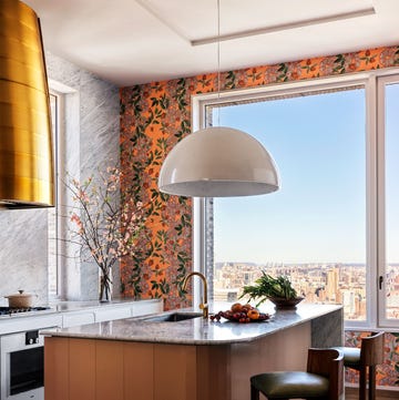 large kitchen window with cityscape view, orange based flowered wallpaper on one wall, marble on wall and countertop, stovetop with brass hood, island with wood base and marble top with sink, large pendant