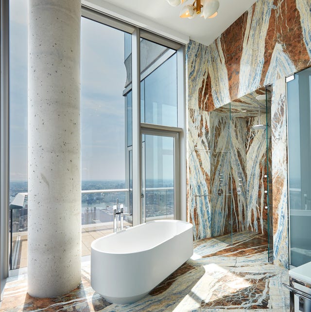 a standalone tub in a bathroom covered in floor to ceiling blue and white and brown marble with a white column and a glass door and window that look out over a very high and far city view