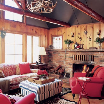 wood panelled living room with large brick fireplace and small mountains animal heads and a red pattenered cushy sofa and matching red leather chairs and a striped blue and white and gray ottoman at center with a tray with flowers and objects on it and antler chandelier above