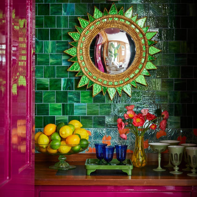 a green sunflower mirror on a green tiled wall in a cubby with bright eggplant magenta laquered drawers and doors and a counter with a bowl of lemons and limes and blue glasses and a yellow vase