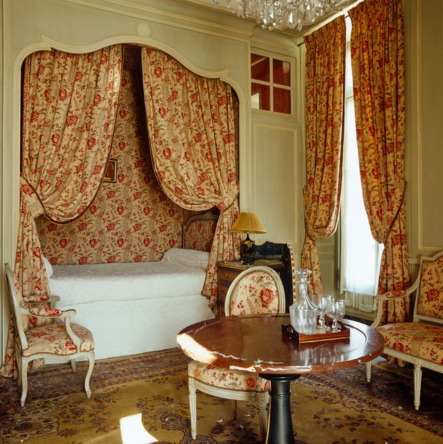 an inset captains bed with red patterned wallpaper behind it and matching curtains and draperies and upholstery on the chairs and sette and a round pedestal table somewhat at centere