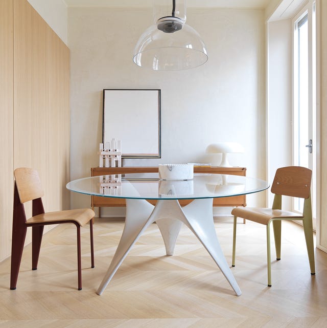 a stark room with light chevron pattern wood floors and a modern round glass top tripod table with wooden chairs around